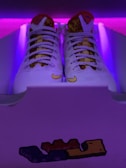 A pair of white sneakers with intricate mesh detailing and a patterned tongue, placed inside a shoe box. The background is illuminated with purple lighting, creating a vibrant contrast. A small graphic featuring block-like shapes is visible on the front of the box.