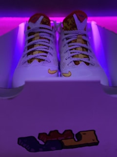 A pair of white sneakers with intricate mesh detailing and a patterned tongue, placed inside a shoe box. The background is illuminated with purple lighting, creating a vibrant contrast. A small graphic featuring block-like shapes is visible on the front of the box.