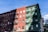 A street view of two adjacent buildings, one painted in red and the other in green, each with multiple floors and fire escape ladders attached to the front. The sky is clear with a few clouds, and a street sign is visible in the foreground promoting a local business.