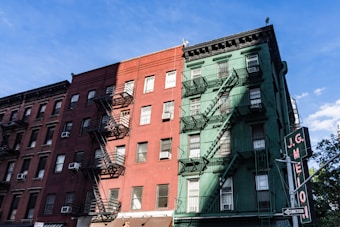 A street view of two adjacent buildings, one painted in red and the other in green, each with multiple floors and fire escape ladders attached to the front. The sky is clear with a few clouds, and a street sign is visible in the foreground promoting a local business.