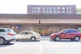 A street scene featuring three parked cars in front of a store named New York Mart. The storefront includes awnings with Chinese characters above it. The background is a red brick building with multiple windows. A small display of produce is visible outside the store, along with a shopping cart. The overall lighting indicates a sunny day.