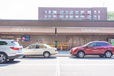 A street scene featuring three parked cars in front of a store named New York Mart. The storefront includes awnings with Chinese characters above it. The background is a red brick building with multiple windows. A small display of produce is visible outside the store, along with a shopping cart. The overall lighting indicates a sunny day.
