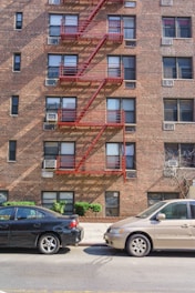 A brick apartment building with multiple windows and red fire escapes is shown. Two cars, one black and the other beige, are parked in front of the building. Green shrubs are planted near the sidewalk, and a tree with bare branches is also visible.