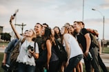 A group of young adults gathers closely together in an outdoor setting, smiling and posing for a selfie taken by one person holding a smartphone. There is a mix of casual clothing, and some people have backpacks or cameras, suggesting an informal and relaxed atmosphere.