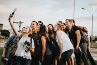 A group of young adults gathers closely together in an outdoor setting, smiling and posing for a selfie taken by one person holding a smartphone. There is a mix of casual clothing, and some people have backpacks or cameras, suggesting an informal and relaxed atmosphere.