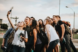 A group of young adults gathers closely together in an outdoor setting, smiling and posing for a selfie taken by one person holding a smartphone. There is a mix of casual clothing, and some people have backpacks or cameras, suggesting an informal and relaxed atmosphere.