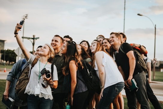 A group of young adults gathers closely together in an outdoor setting, smiling and posing for a selfie taken by one person holding a smartphone. There is a mix of casual clothing, and some people have backpacks or cameras, suggesting an informal and relaxed atmosphere.