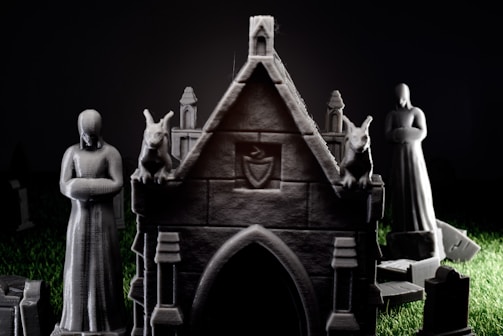 A dimly lit scene features stone-like statuettes in a graveyard setting. In the foreground, a small, ornate mausoleum is flanked by two gargoyle figures. Two human figures stand solemnly, with one slightly obscured in the background. The grass surrounding the structures is an unnatural, vibrant green, contrasting with the dark tones of the statues and grave markers.
