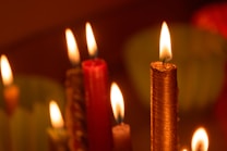 Several lit candles with visible flames create a warm, intimate ambiance. The candles vary in color, with prominent shades of red and gold, and are set against a blurred, soft-focus background.