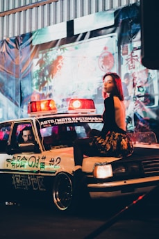 A woman with red hair, dressed in a black top and floral skirt, sits on the hood of a vintage police car with red and blue lights. The car has text and numbers on its side and a man is visible sitting inside. The setting is urban and colorful, with neon lights and signs in the background, creating a vibrant atmosphere.