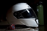 A white motorcycle helmet with a dark visor is placed on a reflective surface. Next to the helmet is a red digital wristwatch. In the background, there is a green water bottle with a logo.