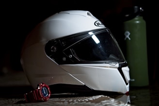 A white motorcycle helmet with a dark visor is placed on a reflective surface. Next to the helmet is a red digital wristwatch. In the background, there is a green water bottle with a logo.