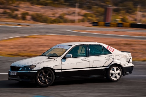 A car with visible wear and tear is drifting on a race track. The vehicle has a white and black color scheme with scuff marks, and there is a 'GDS' sticker on the rear window. The background consists of a blurred outdoor setting, suggesting motion or speed, with dry terrain and barriers.