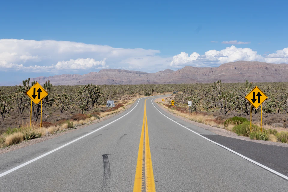 The image depicts a desert road with two yellow diamond-shaped signs on either side, one reading "U" and the other "D". The road is empty, stretching out into the distance towards a mountain range that forms part of the background. The sky above is clear blue with fluffy white clouds.