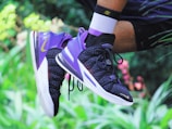 A close-up of a person wearing a pair of mid-air athletic shoes, primarily in purple and black, with visible branding. The background is a blurred natural setting with hints of green foliage and pink flowers. The person is wearing white socks with black accents.