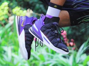 A close-up of a person wearing a pair of mid-air athletic shoes, primarily in purple and black, with visible branding. The background is a blurred natural setting with hints of green foliage and pink flowers. The person is wearing white socks with black accents.