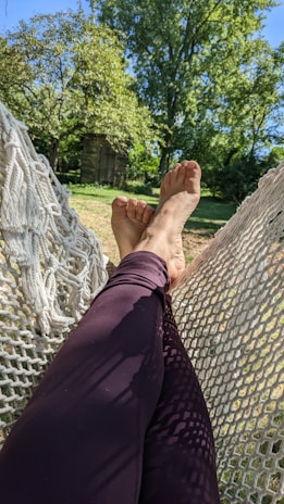 Relaxing in a hammock with crossed legs, surrounded by a lush green garden. Sunlight filters through the leaves, creating dappled shadows on the ground.