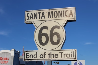 A vintage-style road sign for Route 66 with the words 'Santa Monica' at the top and 'End of the Trail' at the bottom. The background features a clear blue sky and a portion of a building with the 'Bubba Gump' logo visible.