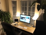 A neatly arranged workspace with a large computer monitor displaying a video platform, accompanied by a smaller tablet, a keyboard, a pair of headphones, and a notepad on a light-colored wooden desk. Two lamps cast a warm glow, and large windows with blinds are visible behind the desk. A large green plant adds a touch of nature to the room.