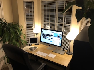 A neatly arranged workspace with a large computer monitor displaying a video platform, accompanied by a smaller tablet, a keyboard, a pair of headphones, and a notepad on a light-colored wooden desk. Two lamps cast a warm glow, and large windows with blinds are visible behind the desk. A large green plant adds a touch of nature to the room.