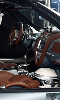 The interior of a luxury car features brown leather seats, a modern ergonomic steering wheel, and a sophisticated dashboard with advanced controls. A racing helmet is placed on the passenger seat beside the gear shift.