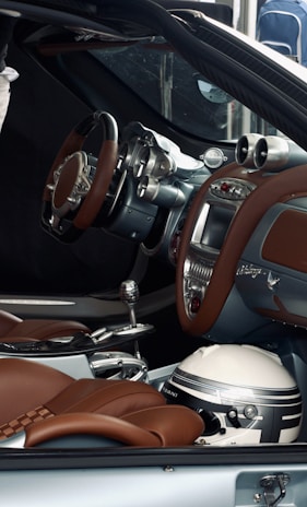 The interior of a luxury car features brown leather seats, a modern ergonomic steering wheel, and a sophisticated dashboard with advanced controls. A racing helmet is placed on the passenger seat beside the gear shift.