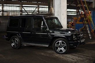 A black SUV is parked in an industrial setting, with concrete pillars and large windows. There is a steel ladder on the right side along with colorful items in the background.