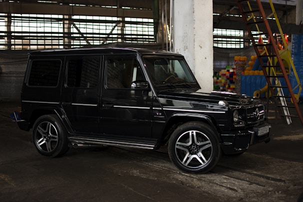 A black SUV is parked in an industrial setting, with concrete pillars and large windows. There is a steel ladder on the right side along with colorful items in the background.