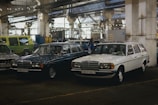 Two vintage Mercedes cars are parked inside an industrial garage or warehouse. The environment has a cluttered, slightly dim atmosphere with visible pipes and beams. Another classic vehicle is partially visible in the background, along with a few people walking around.