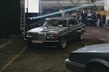 A vintage Mercedes-Benz car is parked indoors in a dimly lit industrial space. The car has a metallic body with a Belarusian license plate. In the background, several people are around equipment, possibly preparing for a photoshoot or event.