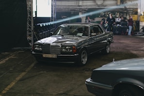 A vintage Mercedes-Benz car is parked indoors in a dimly lit industrial space. The car has a metallic body with a Belarusian license plate. In the background, several people are around equipment, possibly preparing for a photoshoot or event.