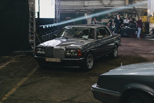 A vintage Mercedes-Benz car is parked indoors in a dimly lit industrial space. The car has a metallic body with a Belarusian license plate. In the background, several people are around equipment, possibly preparing for a photoshoot or event.