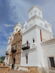 A historic mission-style architectural building with ornate carvings and two distinct towers. The facade is predominately white with intricate stone carvings around the entrance. The sky is bright and clear, contributing to the grandeur of the structure. A few people are gathered near the entrance, adding a sense of scale to the building.