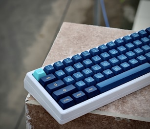 A mechanical keyboard with dark blue and turquoise keycaps is resting on a textured surface. The keyboard features a white case, and the keys display regular QWERTY layout with distinctive coloring.