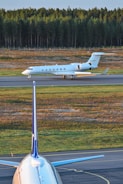 A private jet is taxiing on a runway, with a forested area in the background. In the foreground, the vertical stabilizer and part of the fuselage of a larger commercial airplane are visible. The setting appears to be an airport with a clear sky and natural surroundings.
