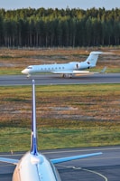 A private jet is taxiing on a runway, with a forested area in the background. In the foreground, the vertical stabilizer and part of the fuselage of a larger commercial airplane are visible. The setting appears to be an airport with a clear sky and natural surroundings.