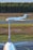 A private jet is taxiing on a runway, with a forested area in the background. In the foreground, the vertical stabilizer and part of the fuselage of a larger commercial airplane are visible. The setting appears to be an airport with a clear sky and natural surroundings.