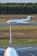 A private jet is taxiing on a runway, with a forested area in the background. In the foreground, the vertical stabilizer and part of the fuselage of a larger commercial airplane are visible. The setting appears to be an airport with a clear sky and natural surroundings.