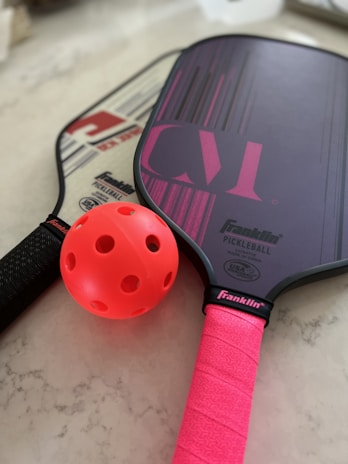 A close-up of two pickleball paddles resting on a light-colored surface. One paddle has a pink grip and design, while the other has a black grip and different branding. A bright pink perforated pickleball is placed between the paddles.