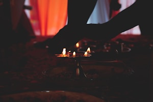 Several small flames are illuminated on traditional brass oil lamps set on a surface. Silhouettes of hands are gently reaching towards the flames as ambient warm light filters through a background curtain.