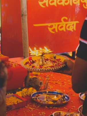 A festive setting with a hand holding a decorative plate containing lit oil lamps and flower petals. The background features bright red fabric with inscriptions in a script language, likely part of a religious or cultural event.
