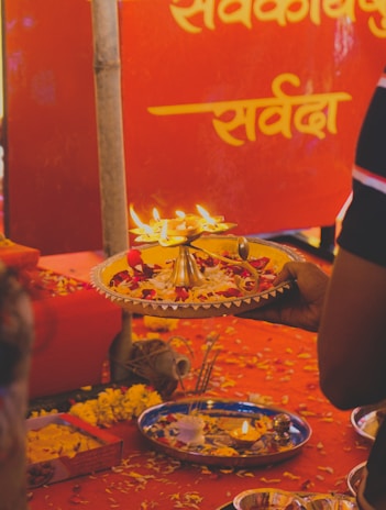 A festive setting with a hand holding a decorative plate containing lit oil lamps and flower petals. The background features bright red fabric with inscriptions in a script language, likely part of a religious or cultural event.