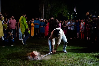 A man is engaging in a traditional game or activity in front of a crowd of onlookers. He is bending down, using a stick or bat to hit an object on the ground. The crowd surrounding him consists of men and women in colorful attire, suggesting a festive or cultural event. Some people are smiling or taking photos, adding to an atmosphere of enjoyment and celebration.