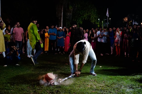 A man is engaging in a traditional game or activity in front of a crowd of onlookers. He is bending down, using a stick or bat to hit an object on the ground. The crowd surrounding him consists of men and women in colorful attire, suggesting a festive or cultural event. Some people are smiling or taking photos, adding to an atmosphere of enjoyment and celebration.