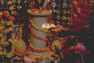A ceremonial setting decorated with garlands of yellow and orange flowers. A hand holding a diya with multiple flames is central, suggesting a religious ritual. A pillar, adorned with flowers, stands in the background. A partially visible figure seems engaged in the ritual.
