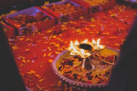 A festive arrangement featuring a brass oil lamp with multiple flames, surrounded by flower petals on a vibrant red textile. In the background, there are boxes containing sweets.