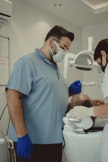 Two dental professionals are attending to a patient seated in a dental chair. The room is equipped with dental instruments and appears clean and clinical. One professional, wearing a mask and blue gloves, stands next to the patient, while another, also masked, works on the patient's head area.