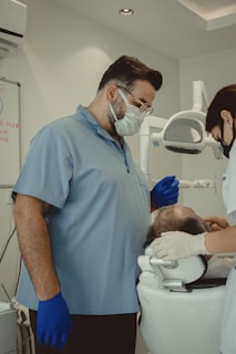 Two dental professionals are attending to a patient seated in a dental chair. The room is equipped with dental instruments and appears clean and clinical. One professional, wearing a mask and blue gloves, stands next to the patient, while another, also masked, works on the patient's head area.