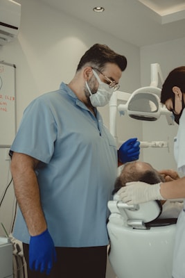 Two dental professionals are attending to a patient seated in a dental chair. The room is equipped with dental instruments and appears clean and clinical. One professional, wearing a mask and blue gloves, stands next to the patient, while another, also masked, works on the patient's head area.