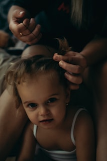 A young child with brown hair is sitting quietly while an adult gently styles their hair, using a small hair tie to create a hairstyle. The setting is intimate and dimly lit, giving a cozy and calm ambience. The child is wearing a white top and looks directly at the camera.